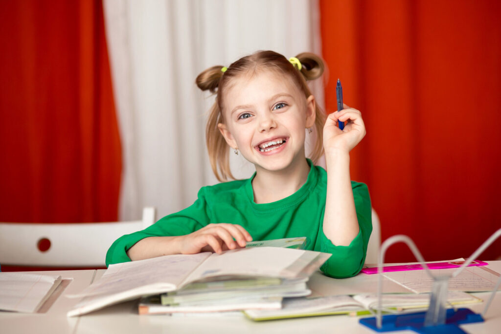 a girl smiling at a table with books and a pen - how to stop homework battles with your child